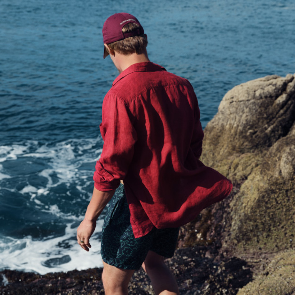 Man walking towards the sea in red linen shirt and swim shorts.