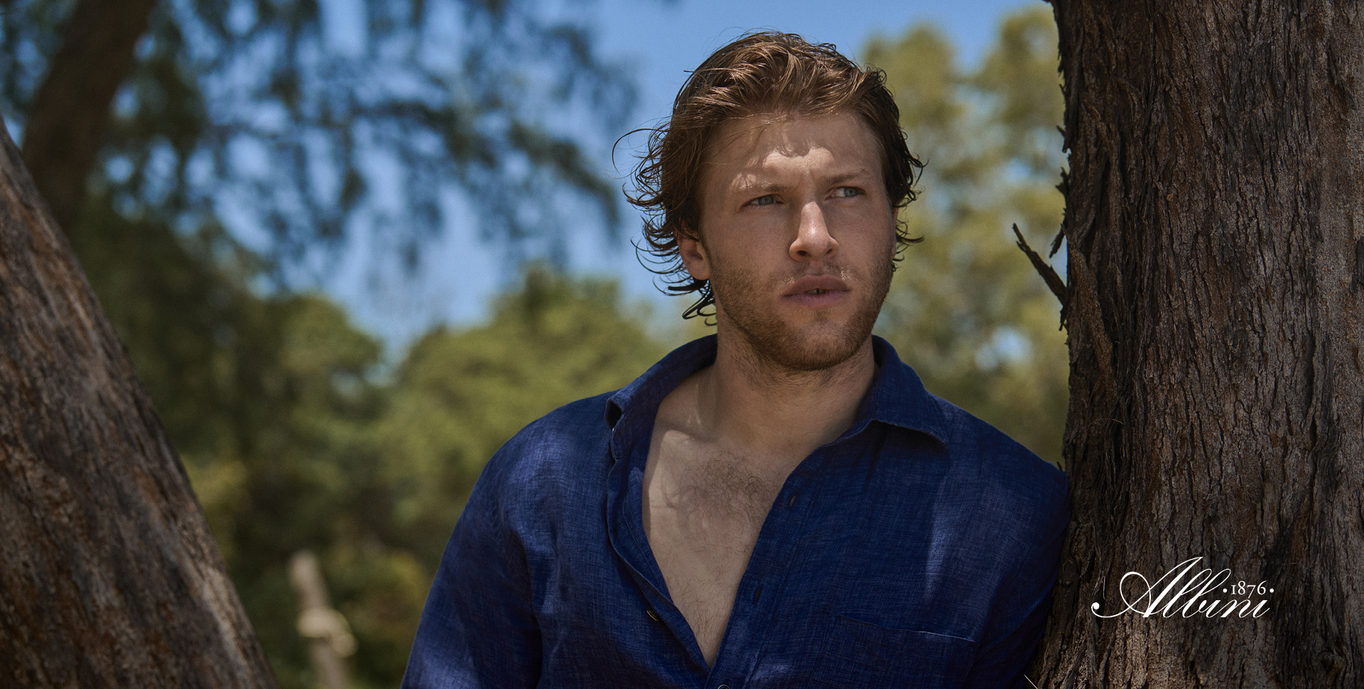 Man leaning against tree wearing dark blue linen shirt.