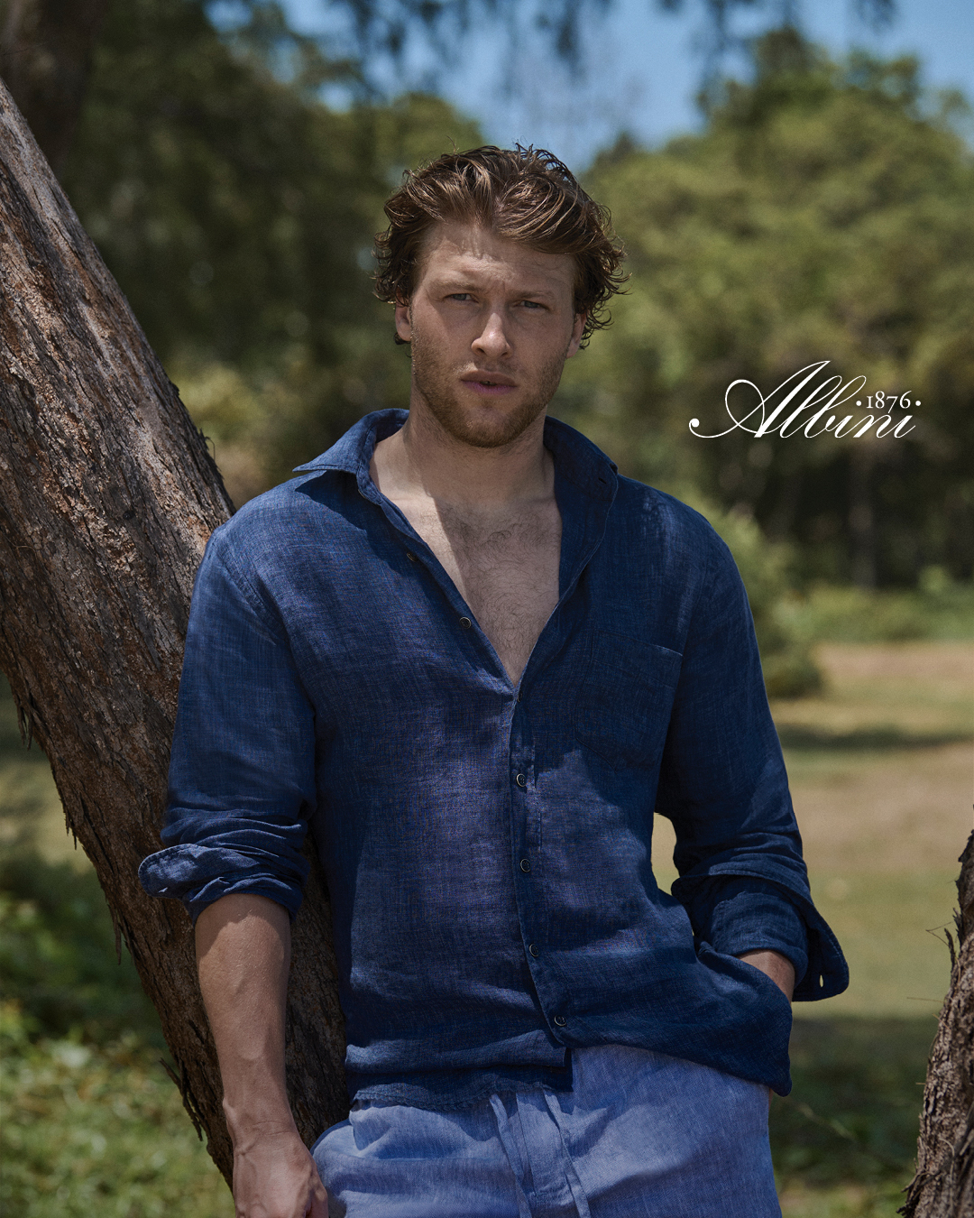 Man leaning against tree wearing dark blue linen shirt.