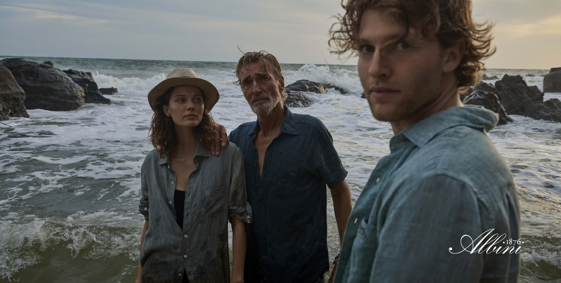 Three people stand together in front of a vast ocean, with waves gently lapping at the shore in the background.
