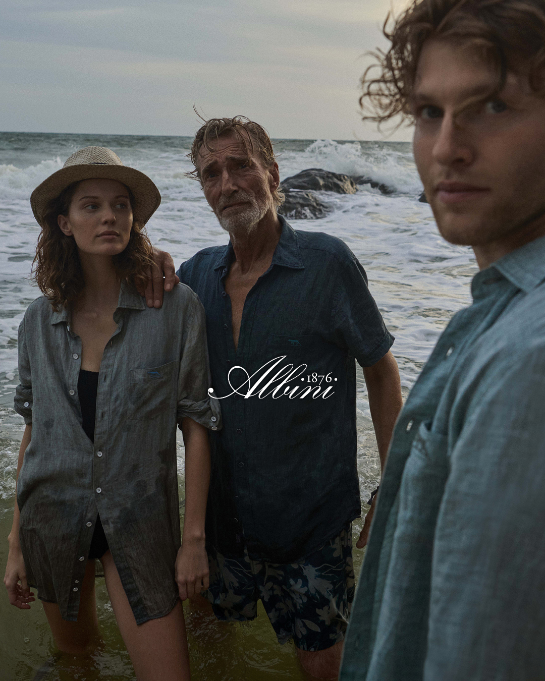 Three people stand together in front of a vast ocean, with waves gently lapping at the shore in the background.