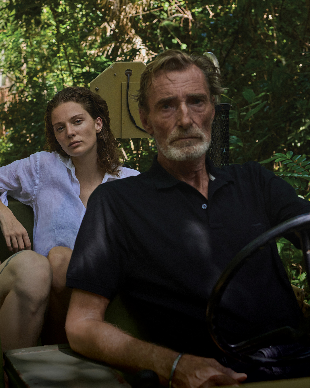 A man driving an open vehicle in a forested area, accompanied by a woman seated beside him and a man slightly behind. The lush green surroundings suggest a natural setting, with trees and trails visible in the background.