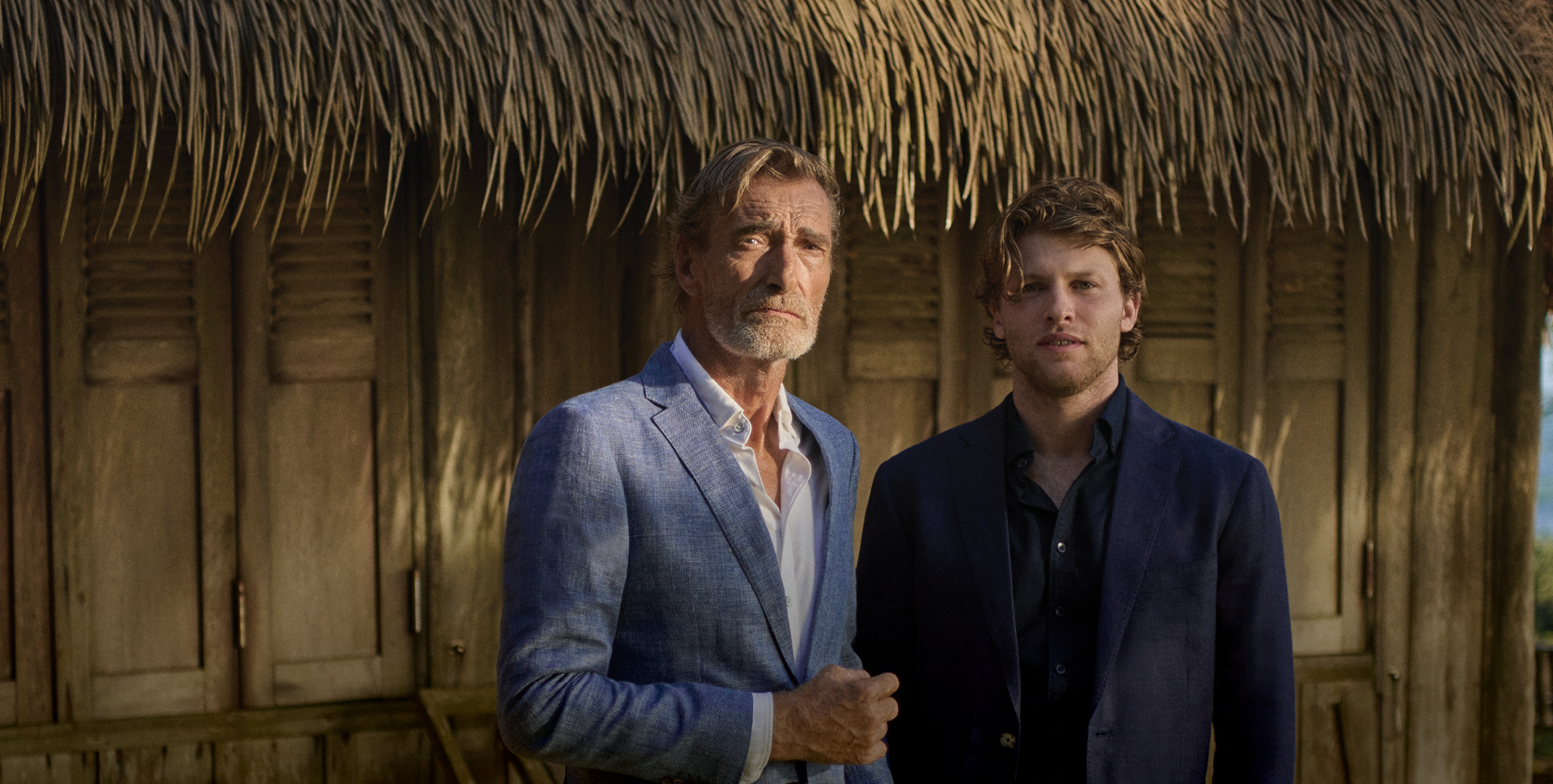 Two men standing in front of a wooden structure with a thatched roof. The older man is wearing a light blue suit jacket over a white shirt, while the younger man is dressed in a dark suit. Both men are looking directly at the camera.
