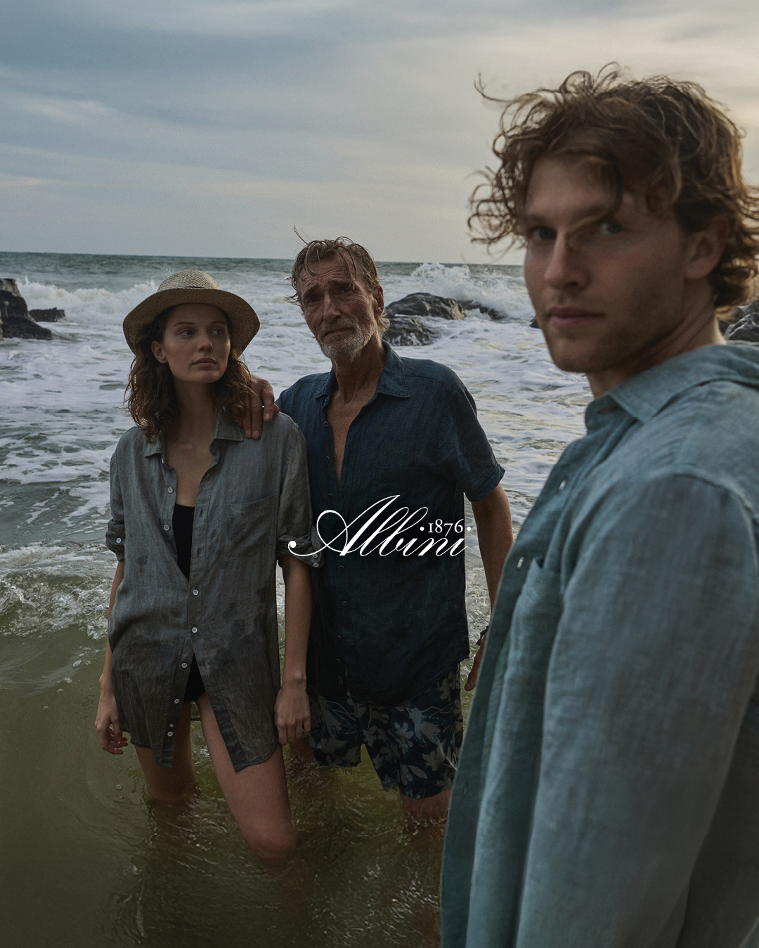 Three people in casual clothing stand in shallow ocean water near rocks at dusk, with waves behind them and a cloudy sky overhead. The word Alltim appears in the lower right corner.
