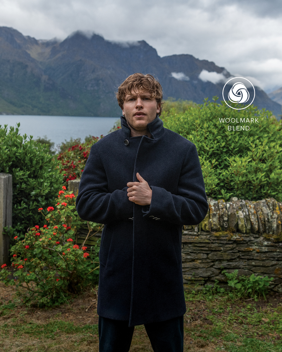 Man stands with lake and stone wall behind him in background, rugged up in a coat