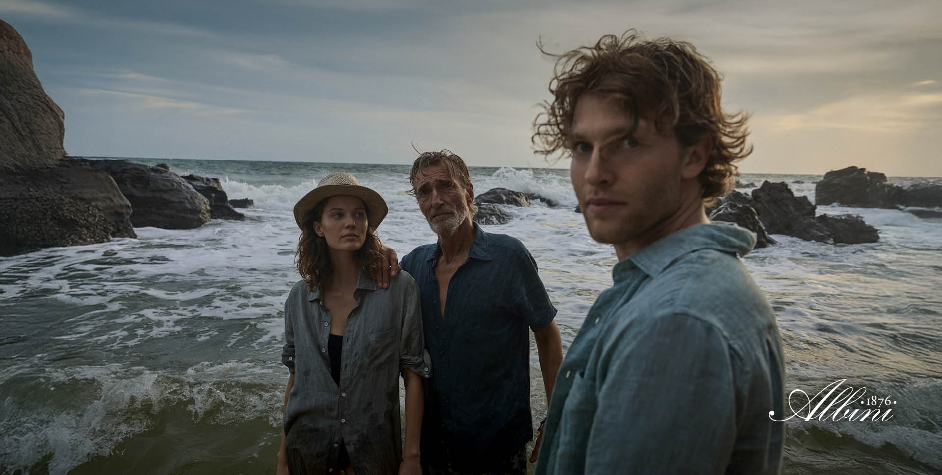 Three people stand in shallow ocean water near rocks at sunset, all wearing blue shirts and looking toward the camera.