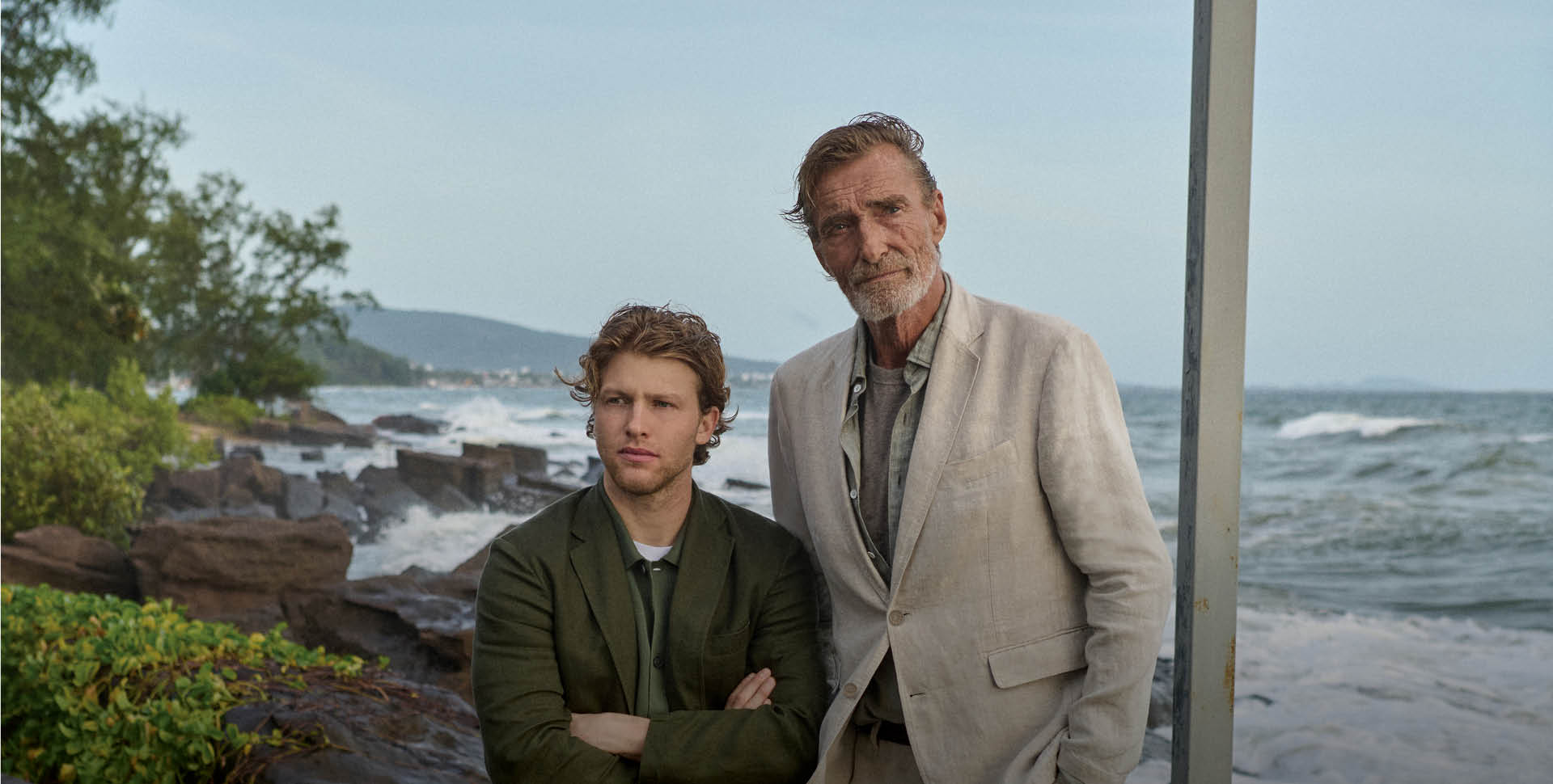 Two men in suits stand by the rocky shoreline, with greenery and waves in the background under an overcast sky.