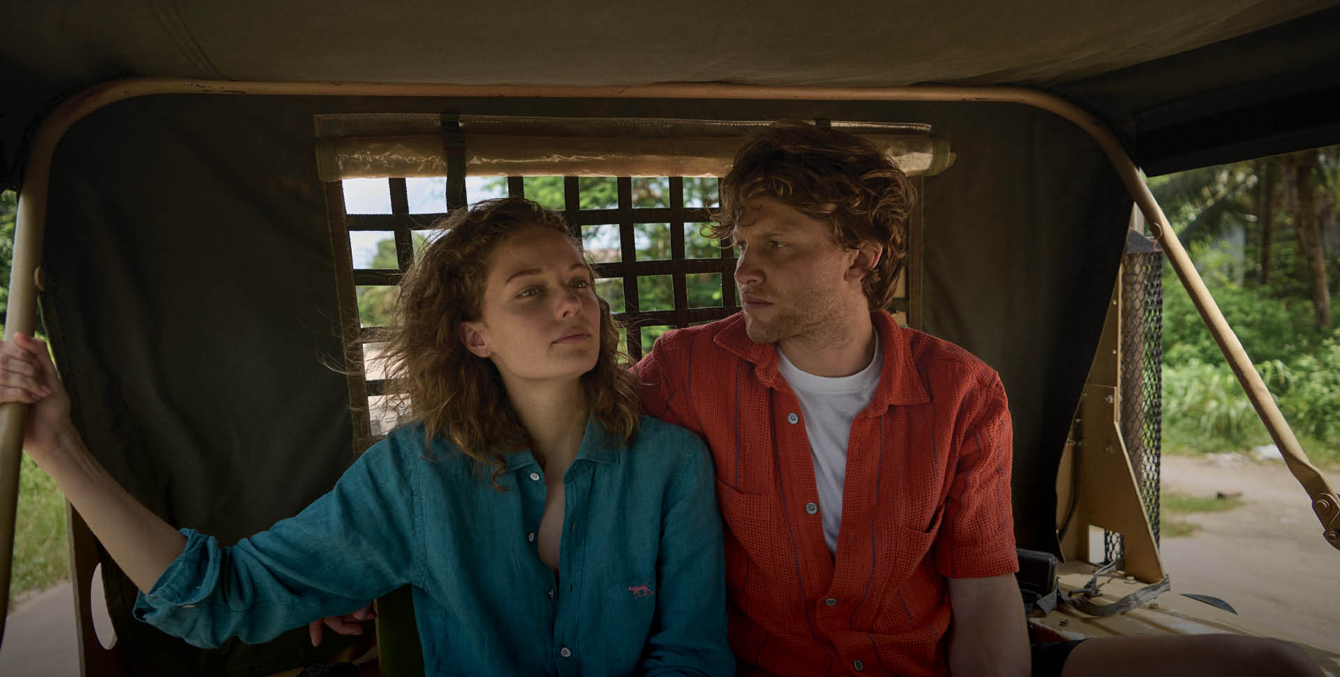 A man and woman sit closely in a moving vehicle, possibly a rickshaw. She wears a teal shirt, looking ahead serenely, while he, in a red shirt, gazes at her. The background shows green foliage, suggesting a tropical setting.