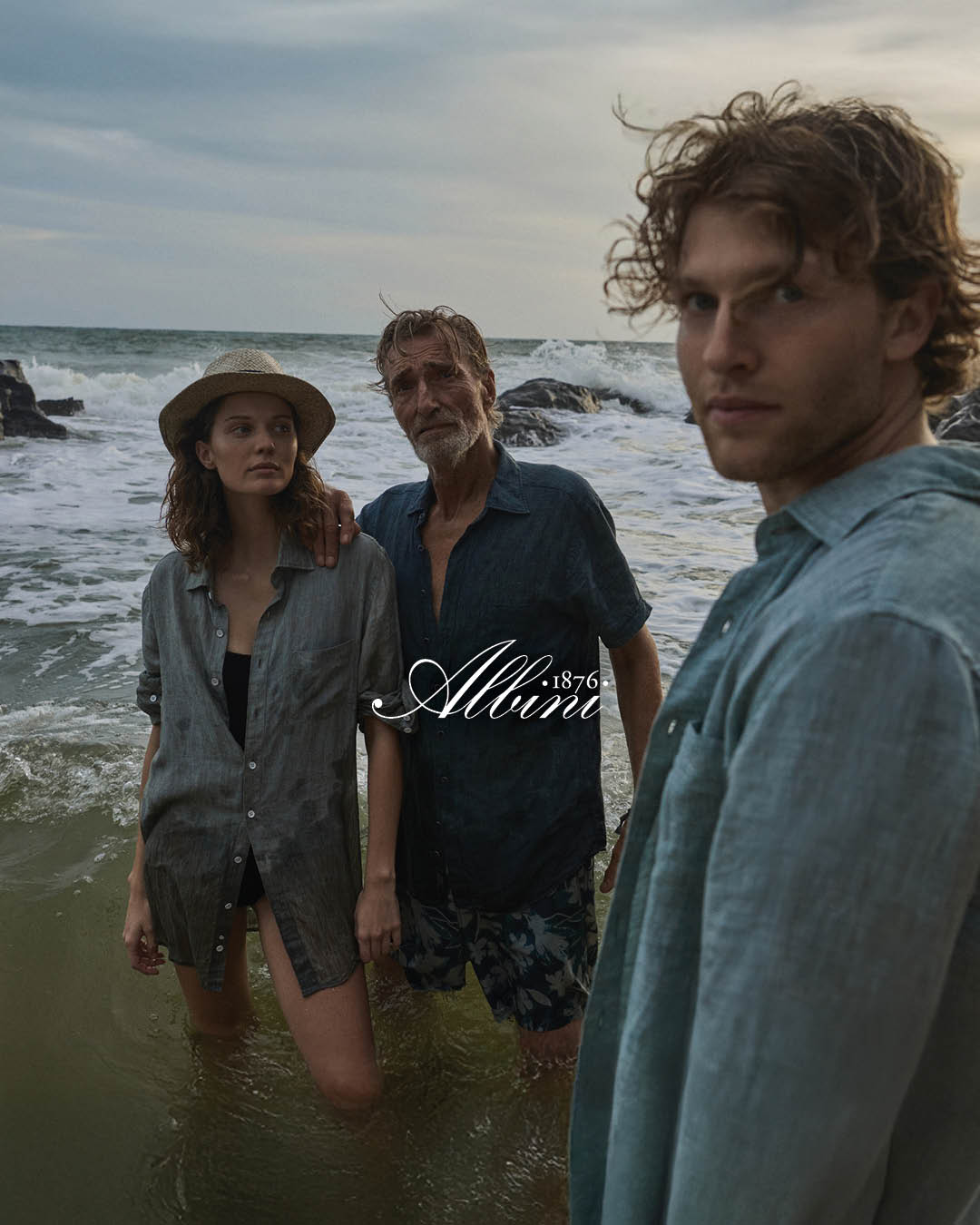 Three people stand in shallow ocean water near rocks at sunset, all wearing blue shirts and looking toward the camera.
