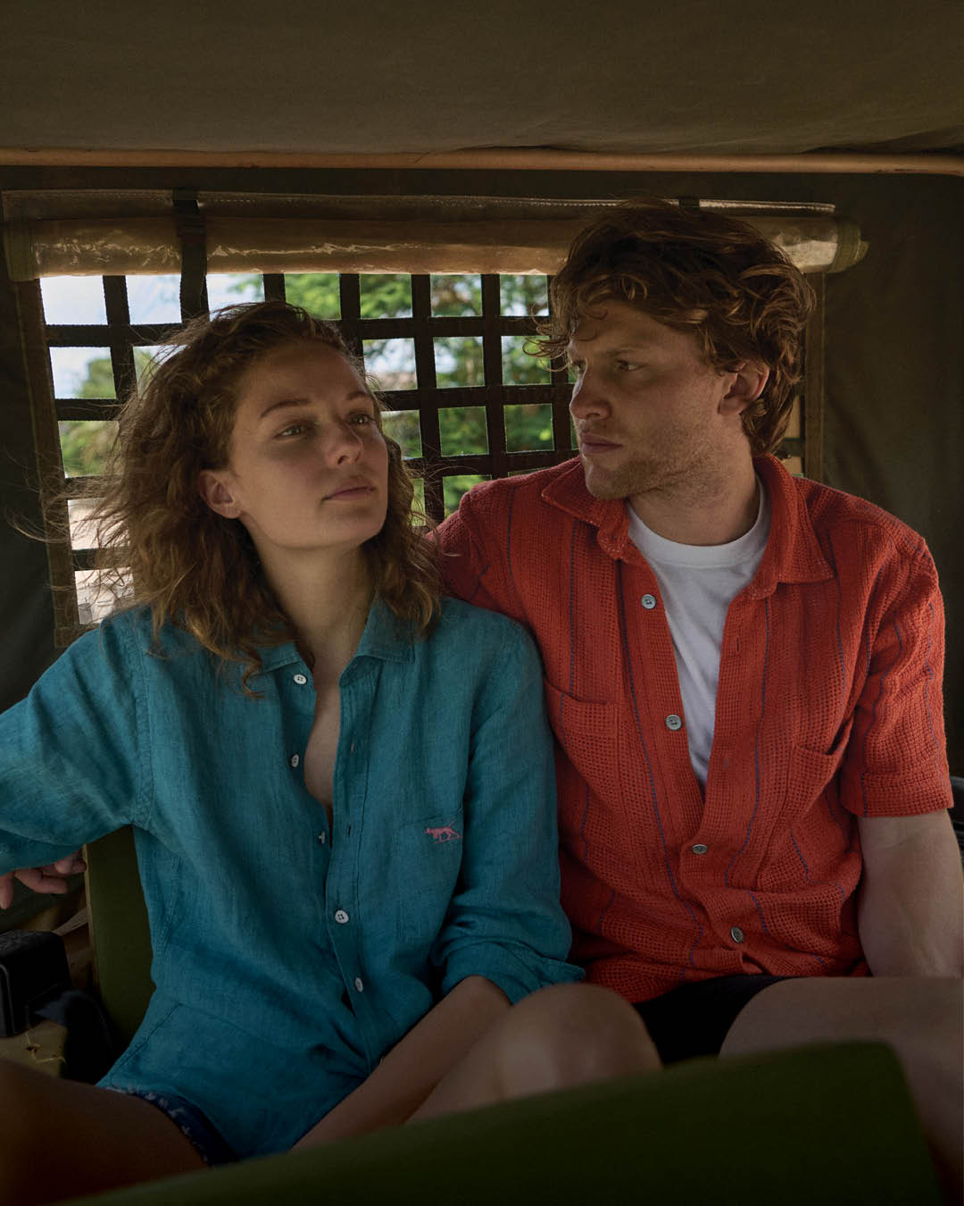 A man and woman sit closely in a moving vehicle, possibly a rickshaw. She wears a teal shirt, looking ahead serenely, while he, in a red shirt, gazes at her. The background shows green foliage, suggesting a tropical setting.