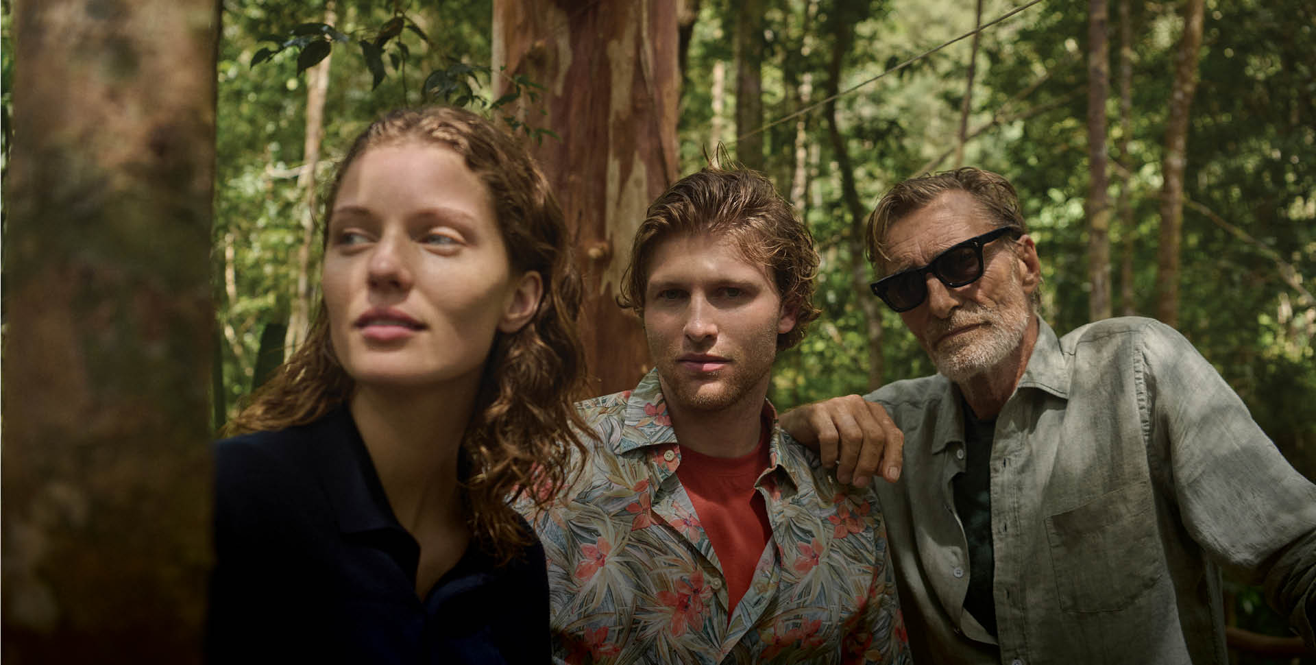 A woman, young man, and older man with sunglasses stand closely together in a forest. They appear thoughtful, surrounded by lush green foliage.