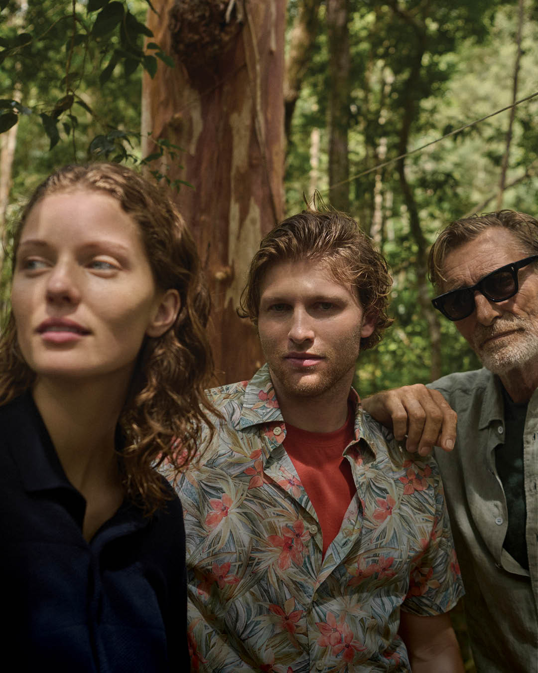 A woman, young man, and older man with sunglasses stand closely together in a forest. They appear thoughtful, surrounded by lush green foliage.