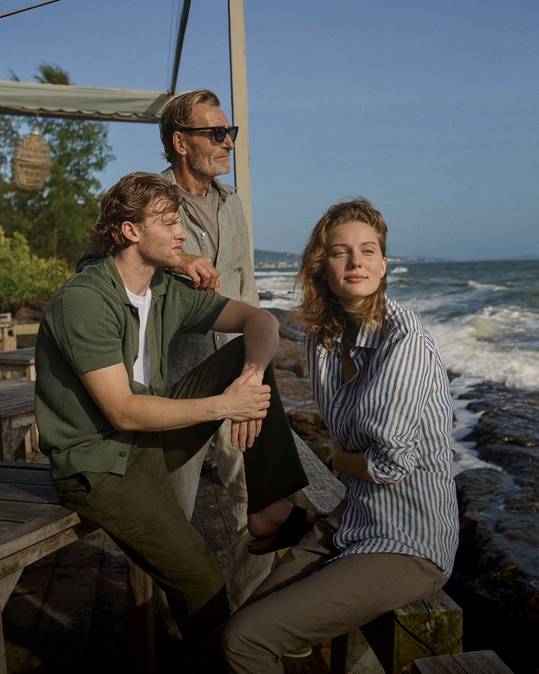 Three individuals standing on a rocky shoreline by the ocean. A man in a green shirt and brown pants sits on a rock, while a woman in a striped shirt and pants stands beside him, looking to the side. An older man in sunglasses stands nearby, gazing out over the water.