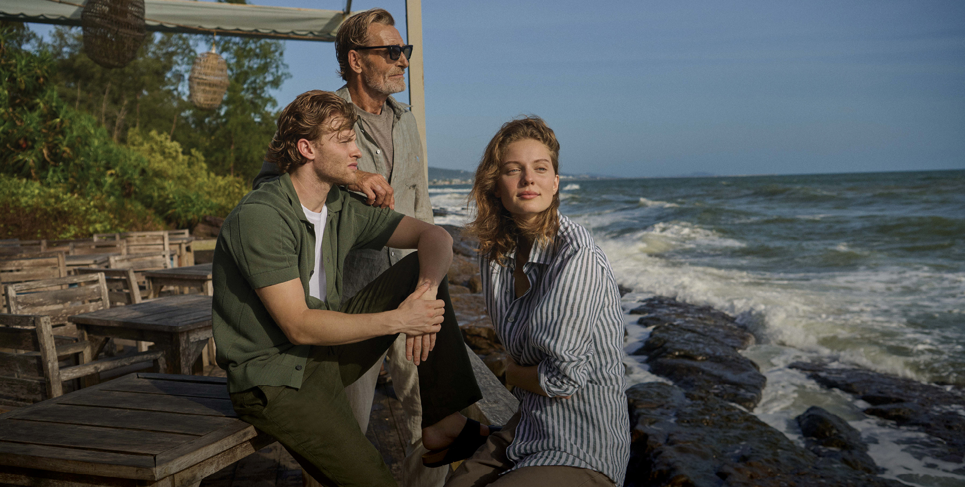 Three individuals standing on a rocky shoreline by the ocean. A man in a green shirt and brown pants sits on a rock, while a woman in a striped shirt and pants stands beside him, looking to the side. An older man in sunglasses stands nearby, gazing out over the water.