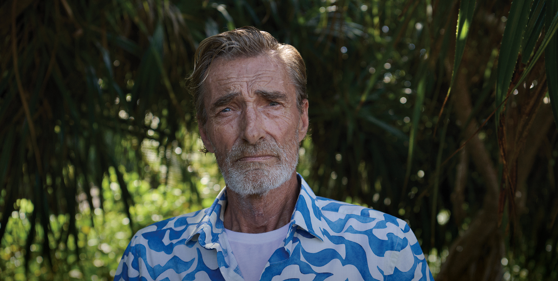 A man with gray hair and a beard stands in front of dense green foliage. He is wearing a blue and white patterned shirt and has a serious expression. The background features palm-like plant leaves that create a natural setting.