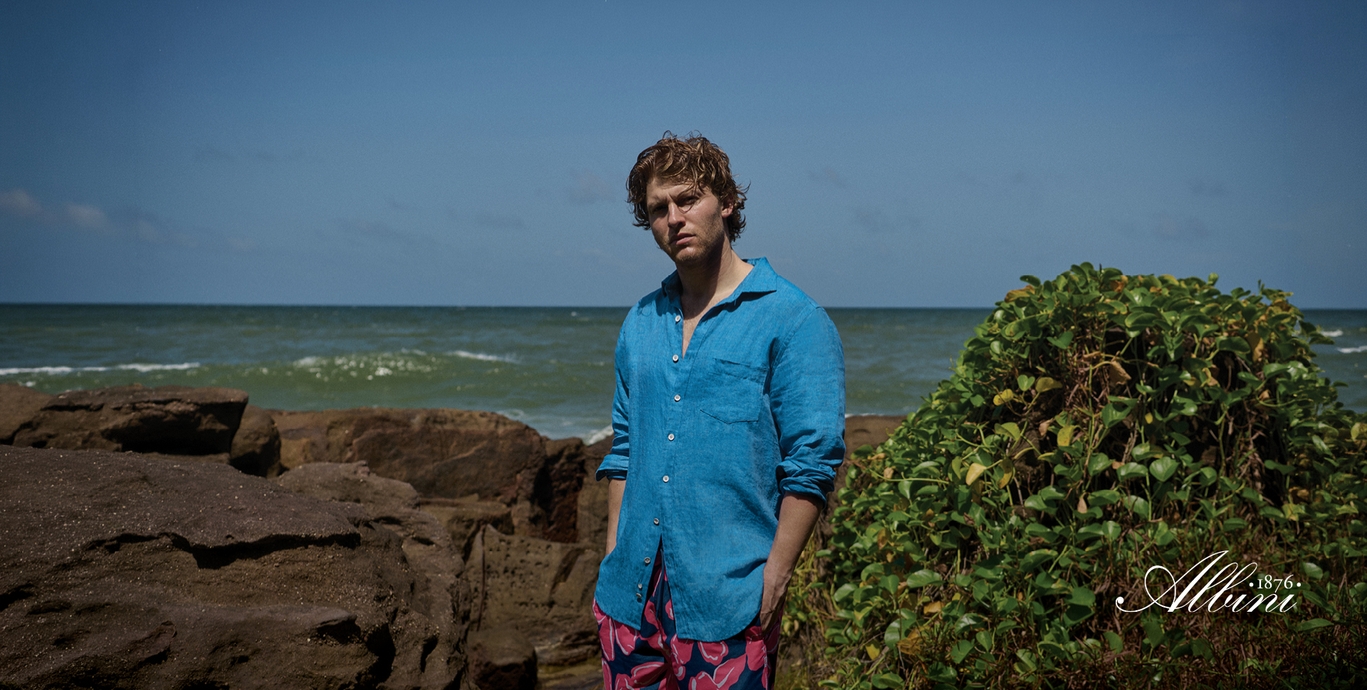 A man standing on rocky shorelines near the ocean, wearing a blue shirt and patterned shorts. He has tousled hair and a neutral expression. In the background, there are waves and greenery, with a clear blue sky.