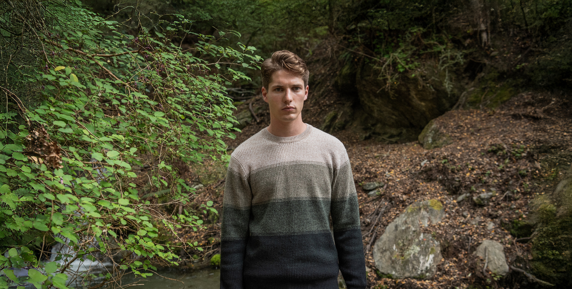 A man in a warm sweater stands in front of a gentle stream, immersed in the beauty of nature.