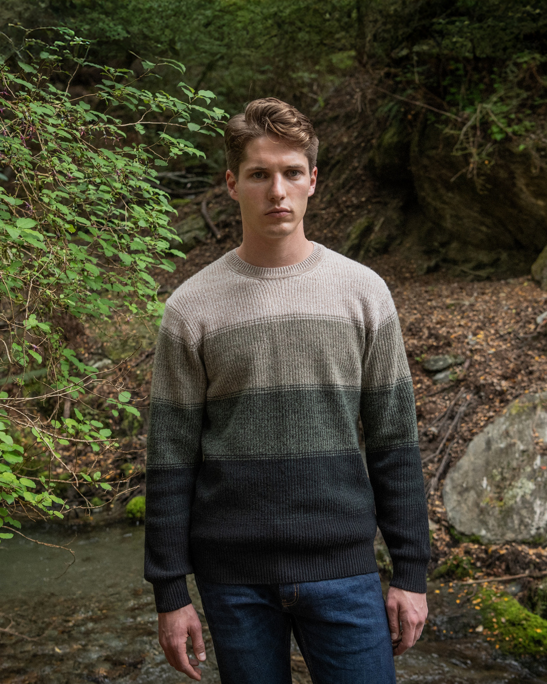 A man in a warm sweater stands in front of a gentle stream, immersed in the beauty of nature.
